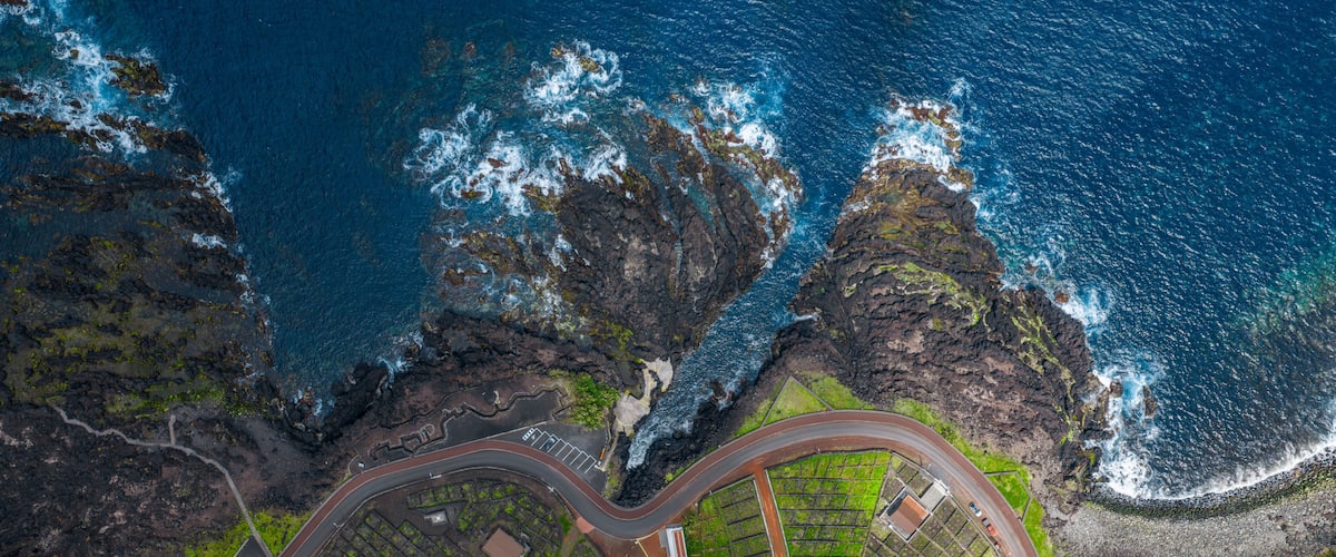 Aerial view of Piscinas Naturais Biscoitos featuring volcanic rock formations, a winding coastal road, and lush green vineyards in Biscoitos, Azores, Portugal.