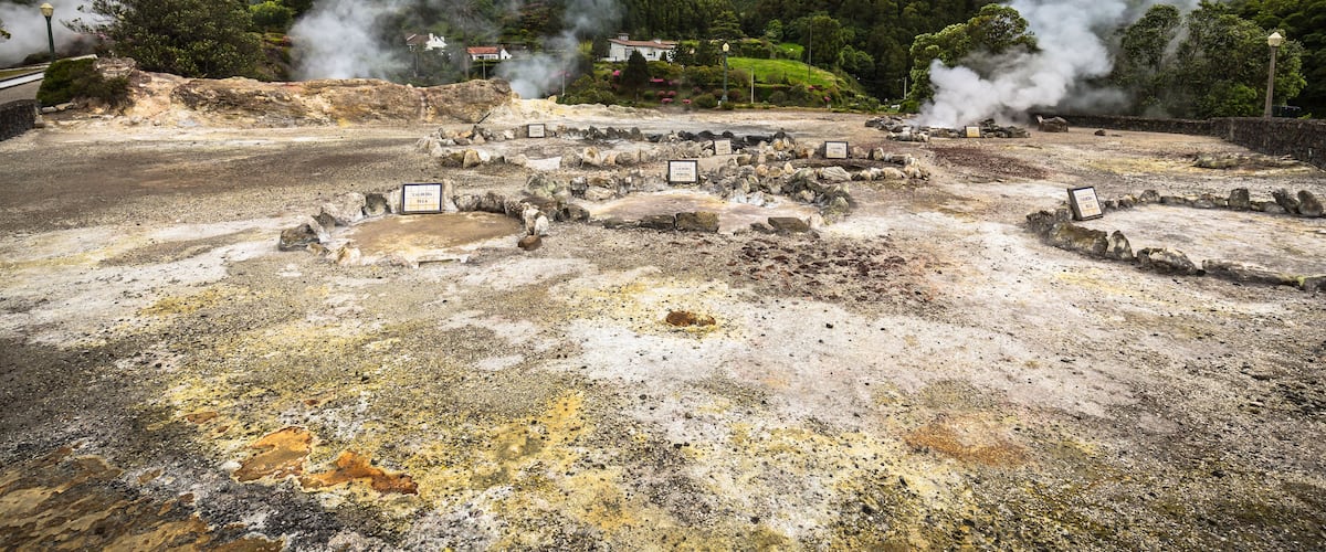 A geothermal area with steam vents, rocky terrain, and a backdrop of green hills.