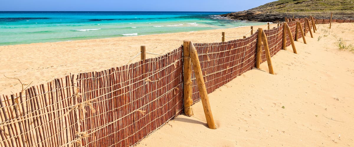 A beach with a bamboo fence, clear turquoise water, and a clear sky.
