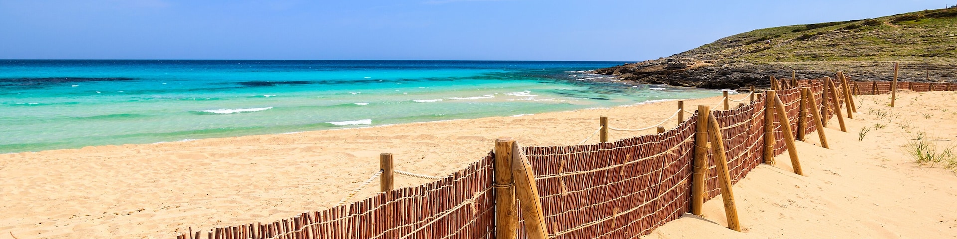 A beach with a bamboo fence, clear turquoise water, and a clear sky.