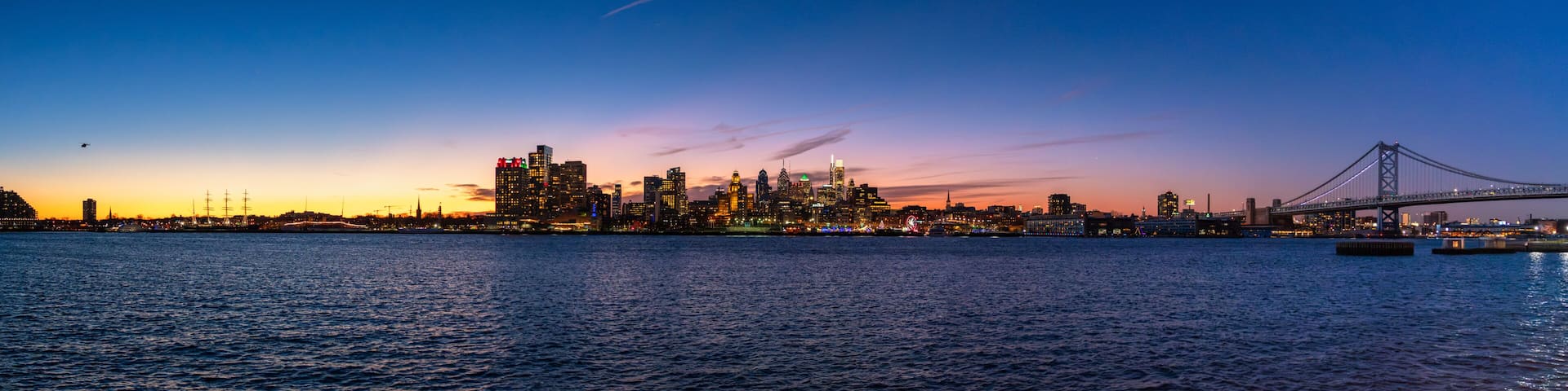 A city skyline at dusk with illuminated buildings and a bridge over water.