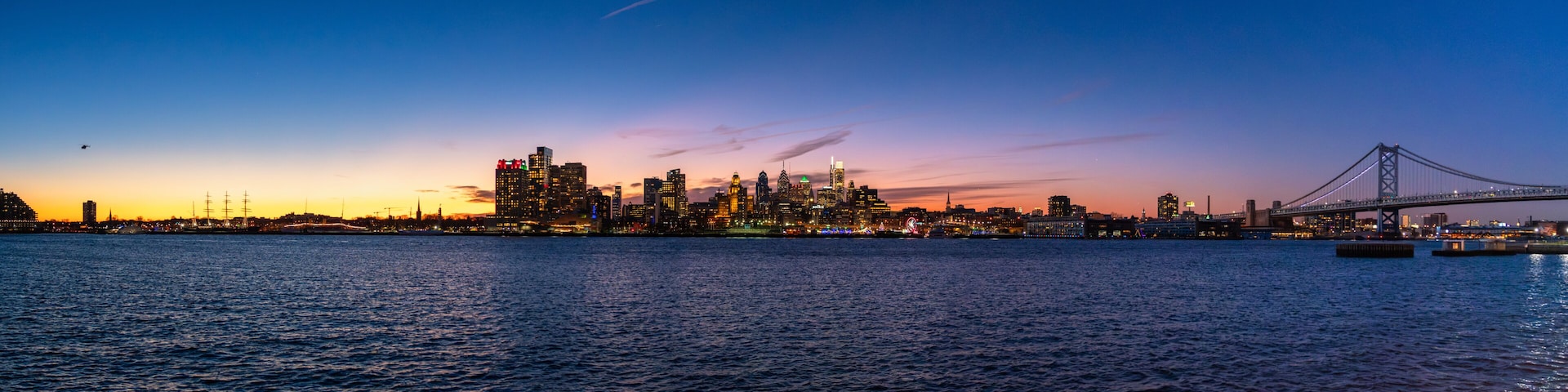 A city skyline at dusk with illuminated buildings and a bridge over water.