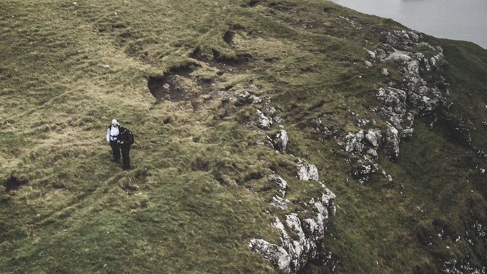 A person hiking on a grassy hillside with rocky outcrops and a large rock formation in the distance.