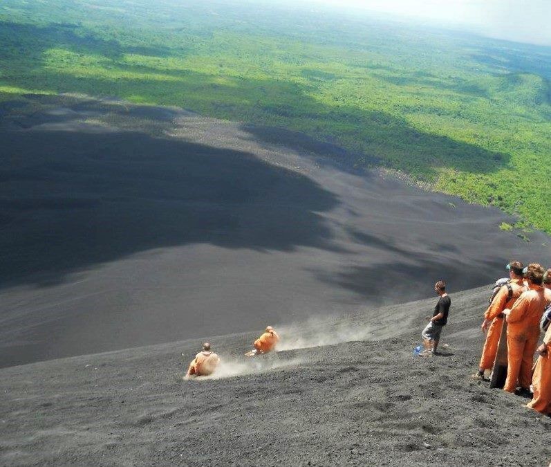 Paisaje volcánico con un cráter grande y personas con trajes protectores naranja observando.