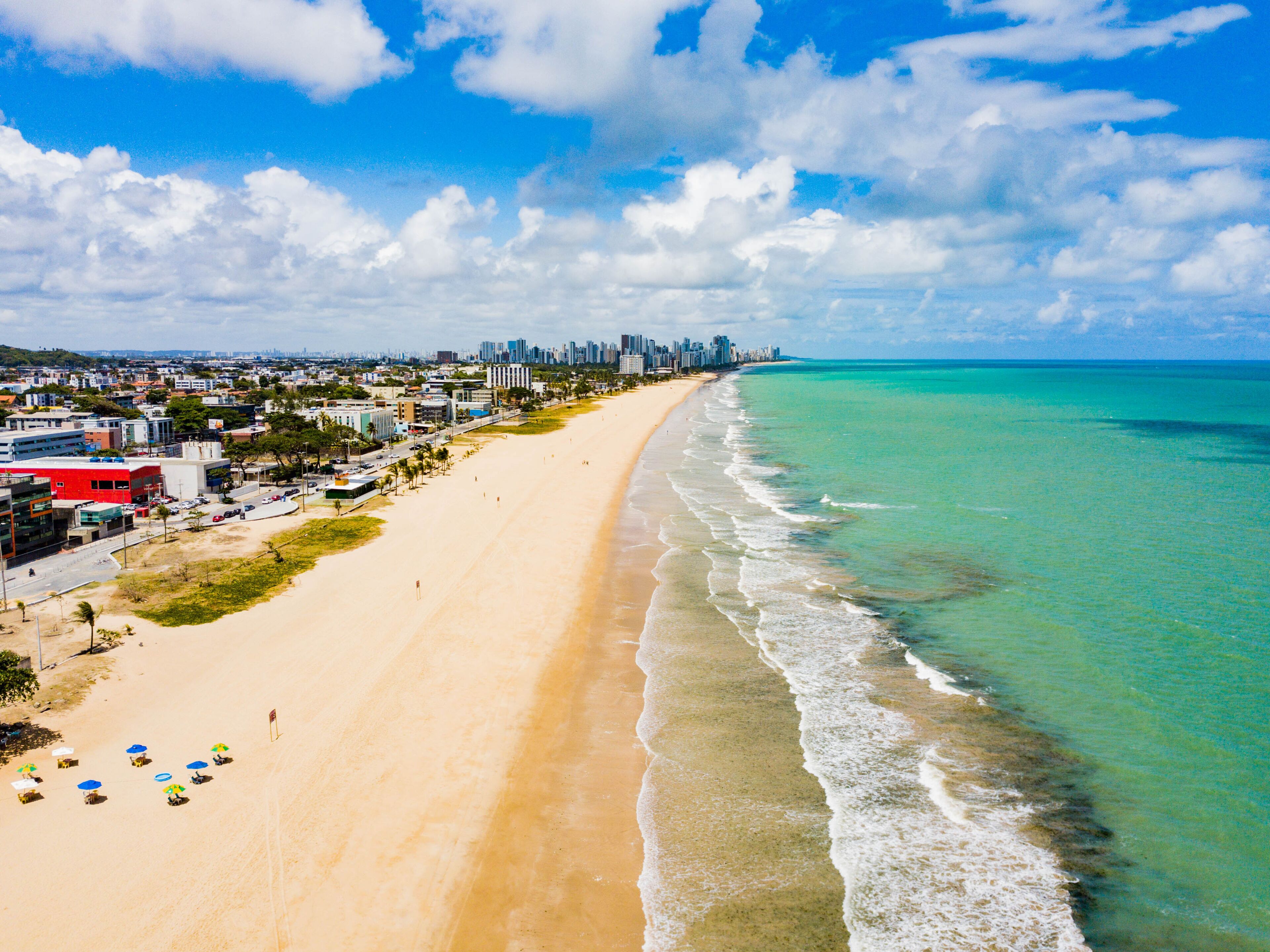 A wide sandy beach with clear turquoise waters, a city skyline in the distance, and a few beach umbrellas.