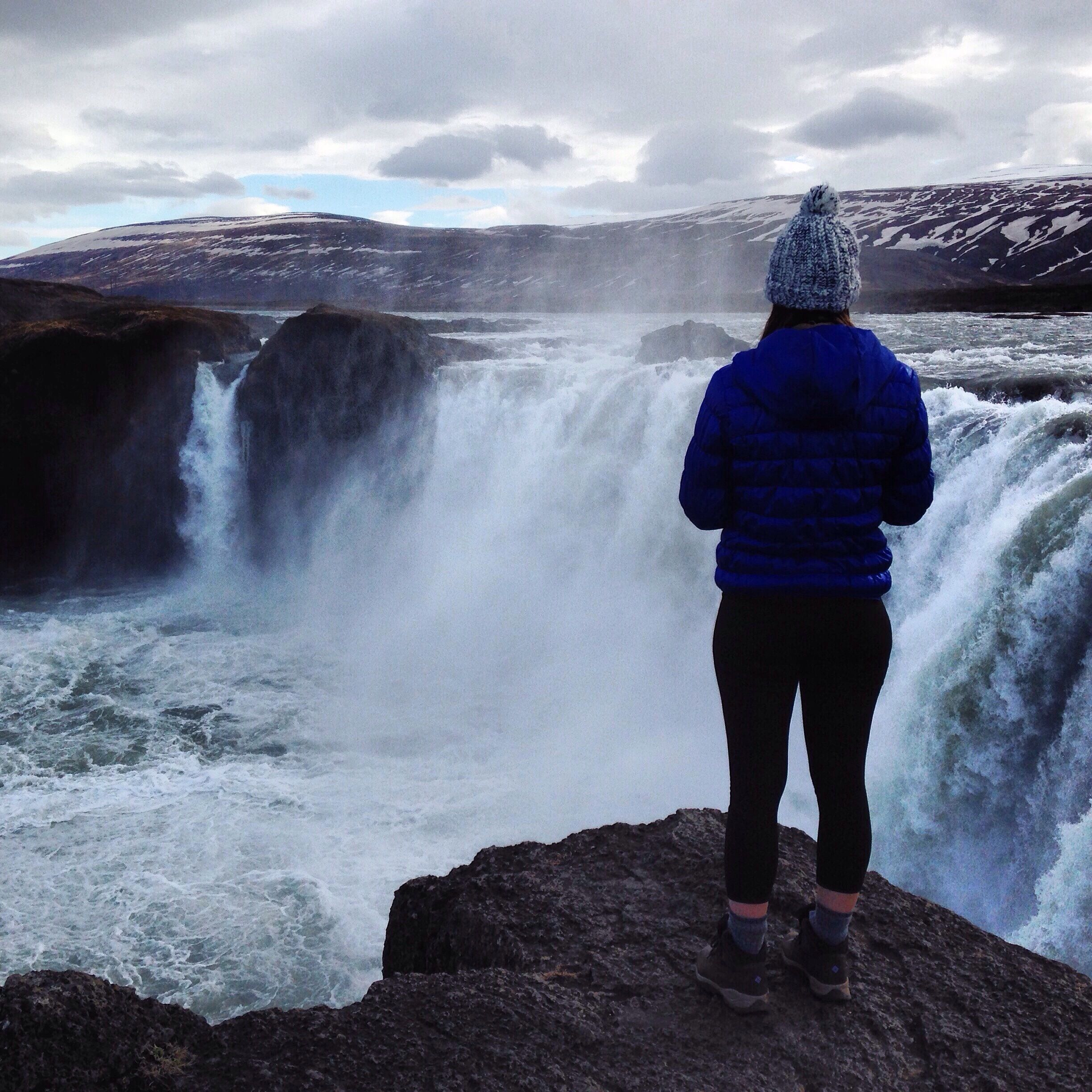 A person standing on a rock, overlooking a powerful waterfall in a mountainous landscape.