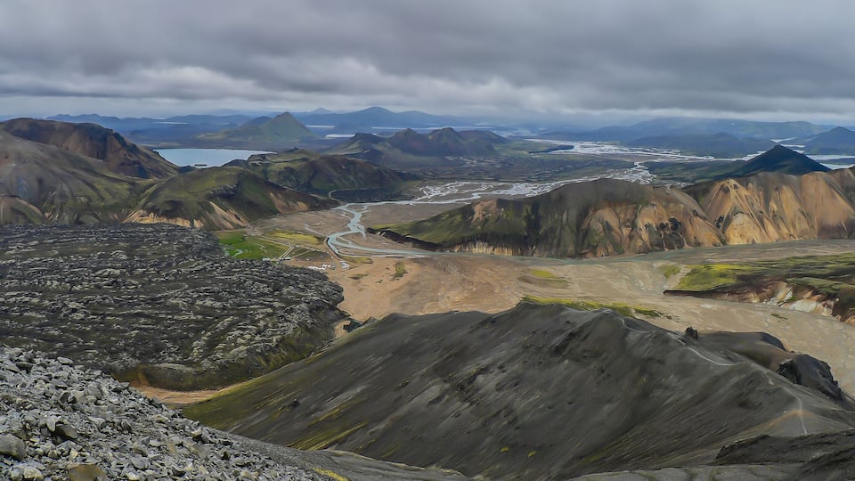 A rugged mountainous landscape with a winding river and distant mountains under a cloudy sky.