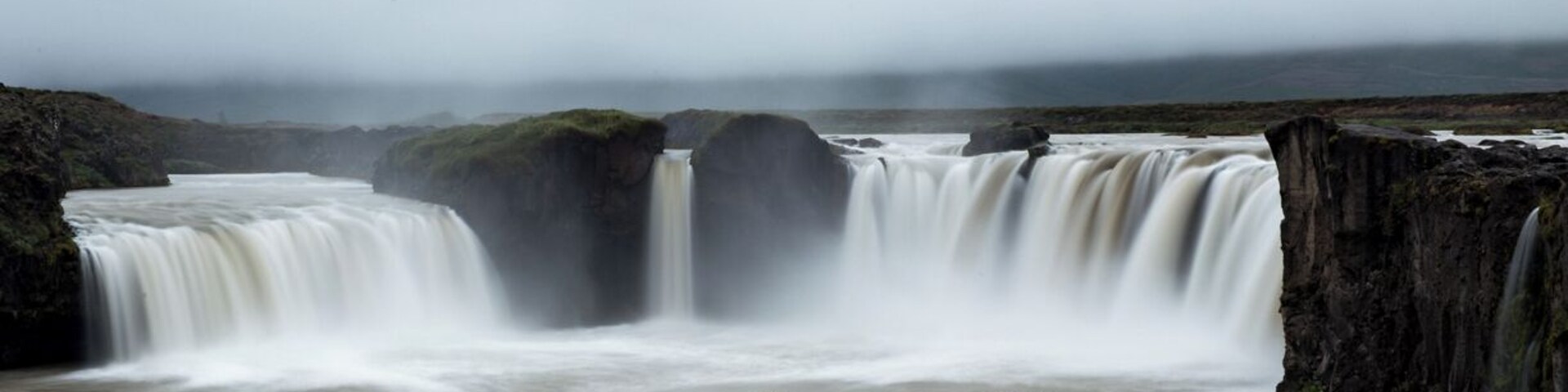 Uma cascata desce sobre rochas em uma paisagem enevoada.