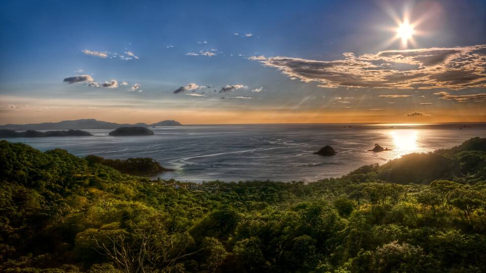 Un paysage côtier caractérisé par une végétation luxuriante, un ciel dégagé et le soleil se couchant sur l’eau.