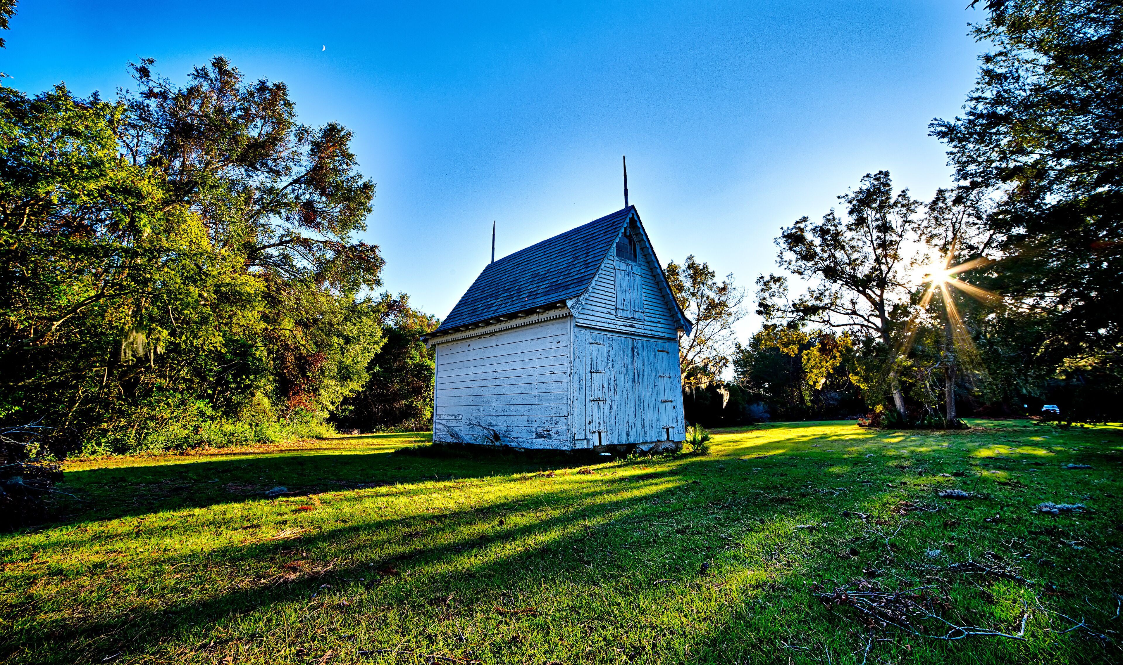 A small white wooden building with a blue roof, surrounded by lush green trees and grass under a clear blue sky.