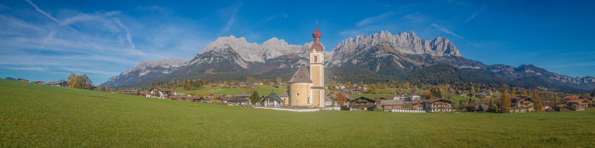 A church with a red dome and a bell tower, surrounded by a village and mountains.