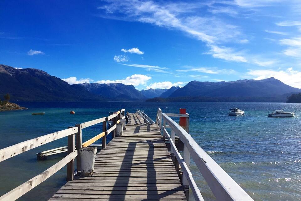 Un muelle de madera que se adentra en un lago con barcos, rodeado de montañas.