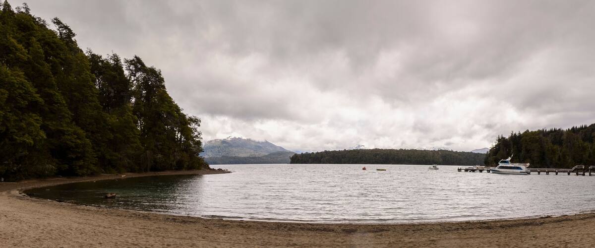 A beach with a pier extending into calm waters, surrounded by dense forests and mountains in the background.