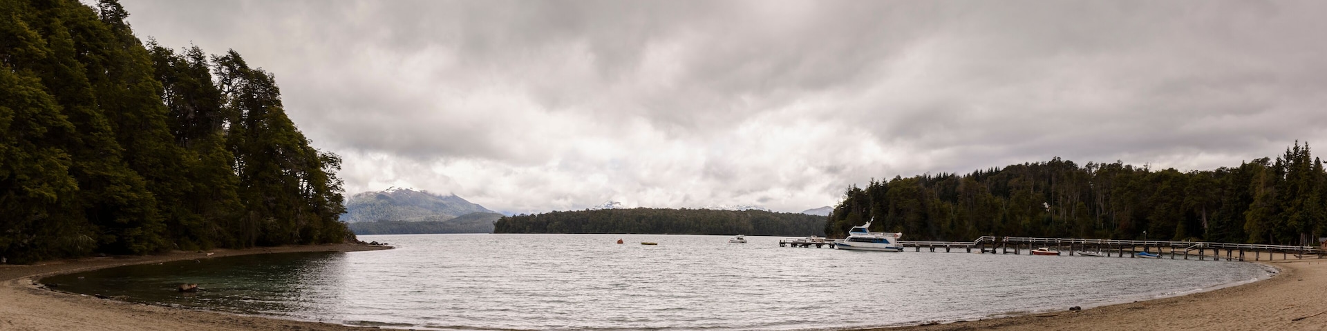 A beach with a pier extending into calm waters, surrounded by dense forests and mountains in the background.