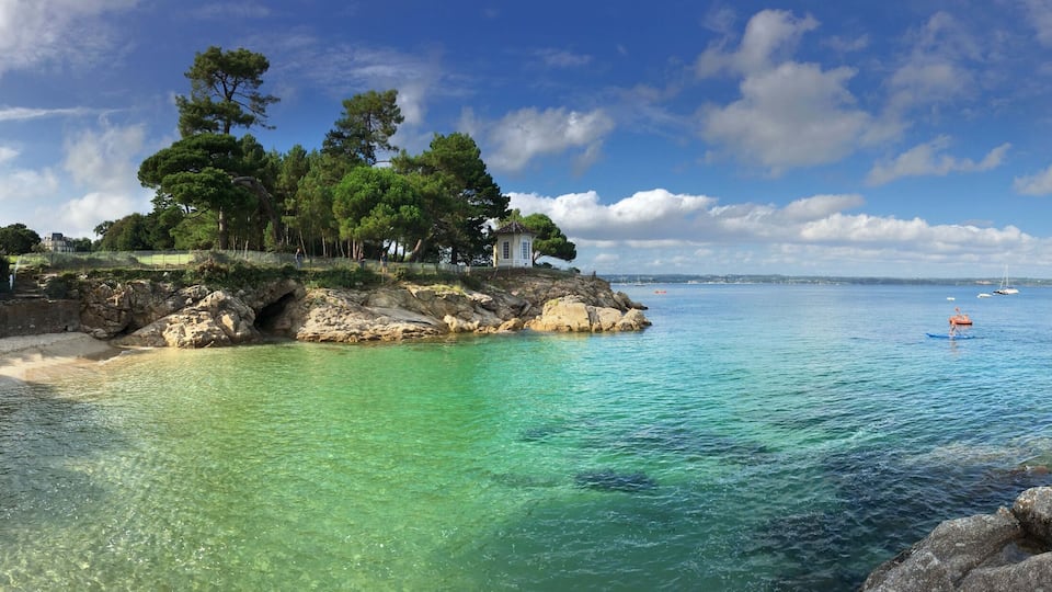 A coastal landscape with clear turquoise waters, rocky outcrops, and a small white building surrounded by trees.