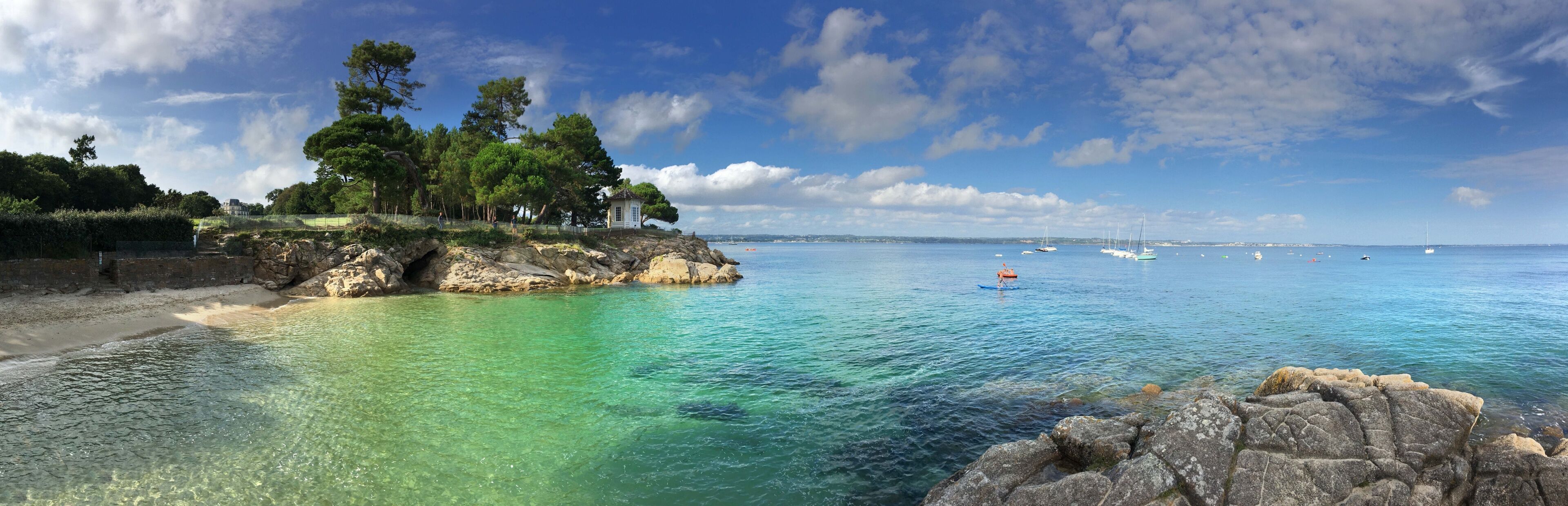 A coastal landscape with clear turquoise waters, rocky outcrops, and a small white building surrounded by trees.