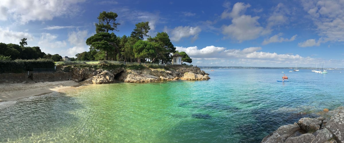 A coastal landscape with clear turquoise waters, rocky outcrops, and a small white building surrounded by trees.