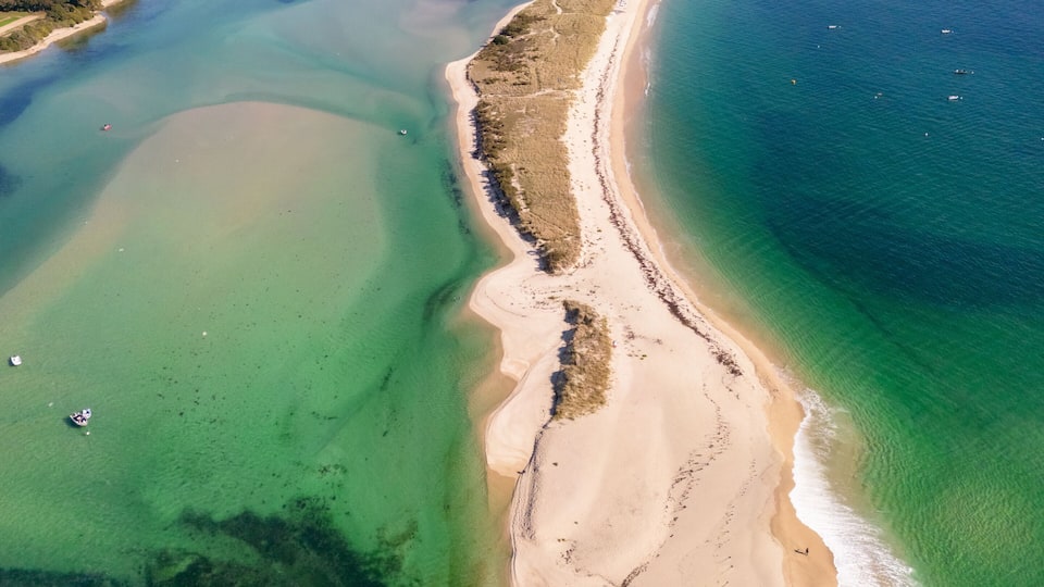 An aerial view of a coastal area with a sandy beach, clear blue waters, and a distant shoreline with buildings.