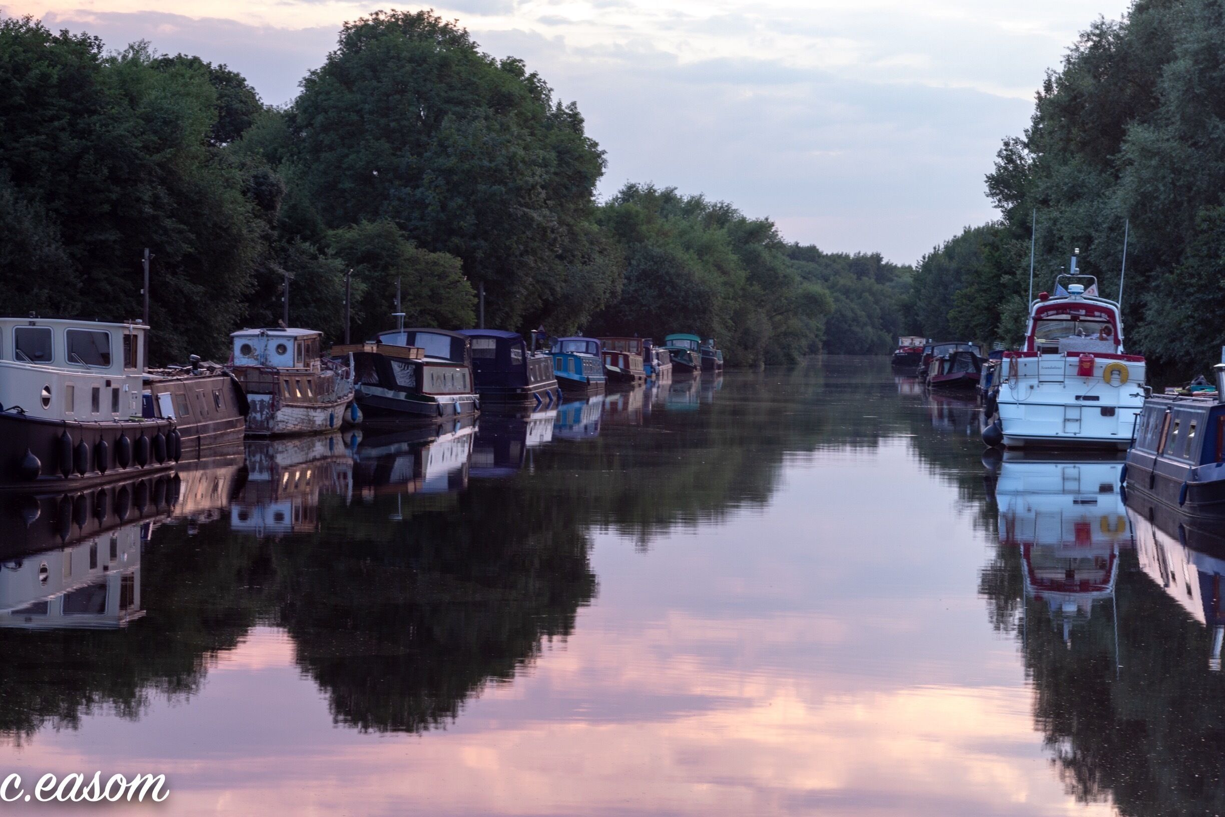A canal lined with boats and trees reflecting on the water.