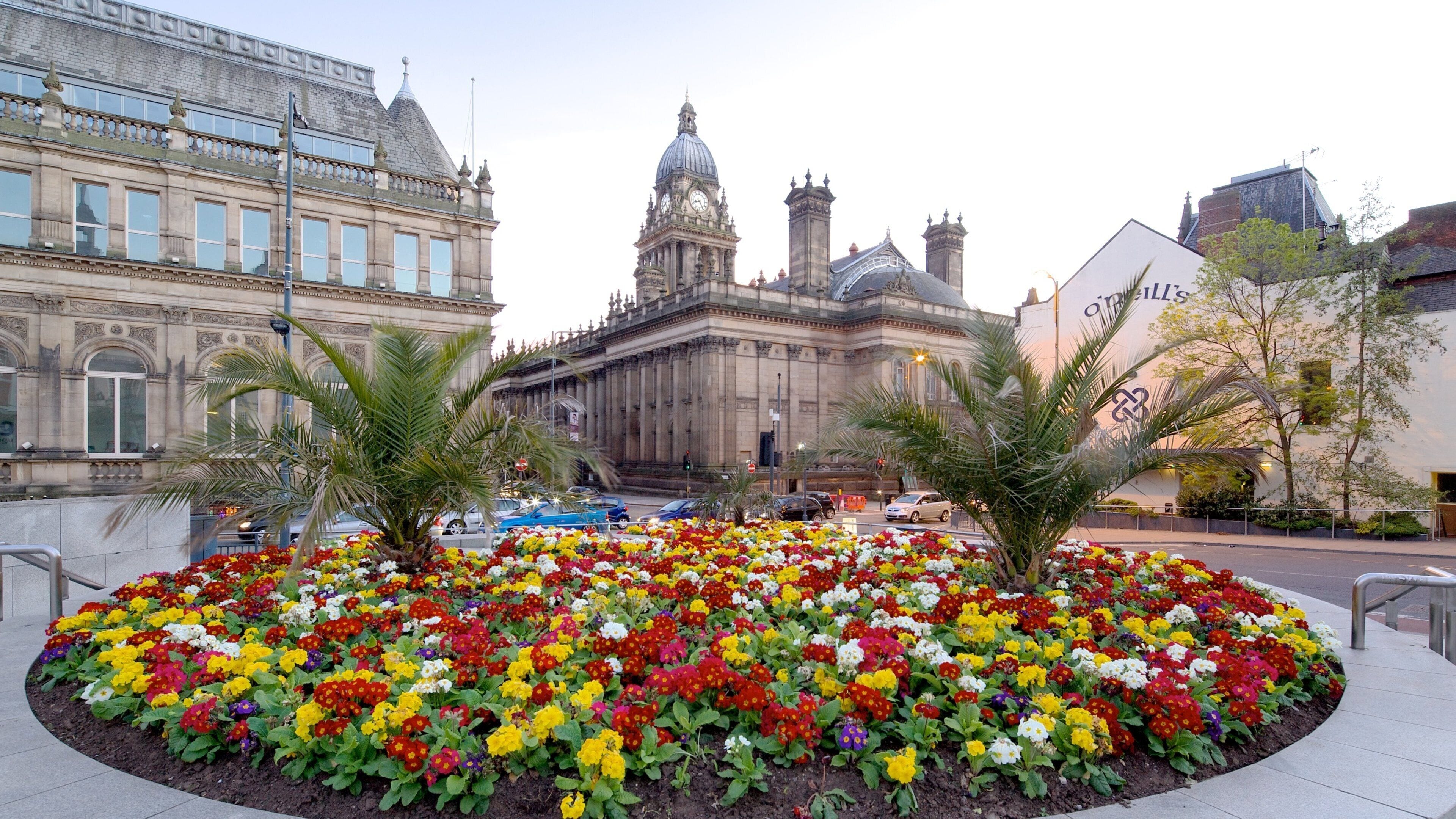 A historic building with a clock tower, palm trees, and a flower bed in the foreground.