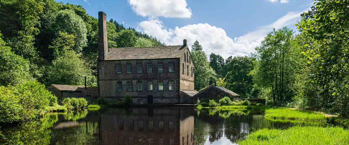 Um edifício histórico de pedra com uma chaminé grande, refletido em um rio calmo rodeado por vegetação exuberante.