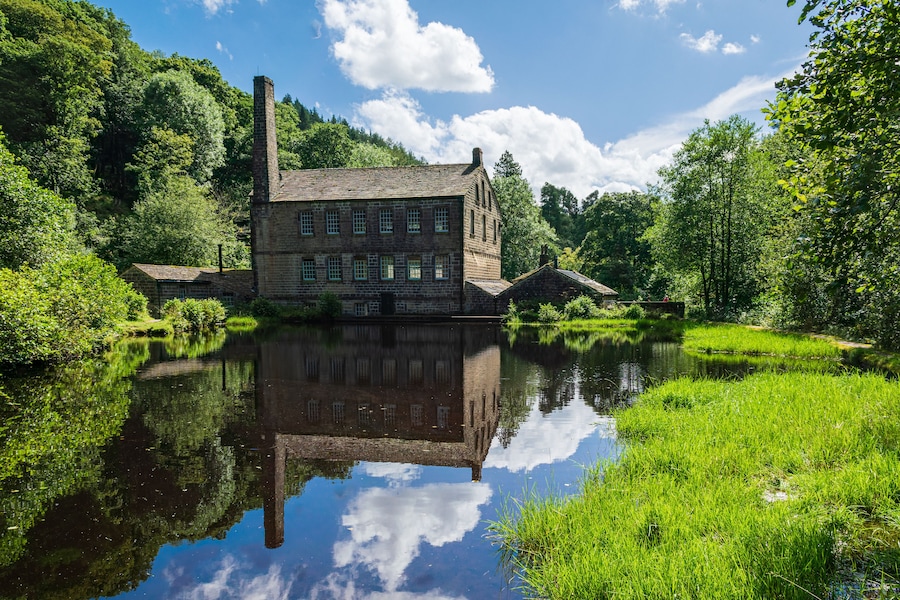 En historisk bygning i sten med en stor skorsten, der spejles i en rolig flod omgivet af frodig vegetation.