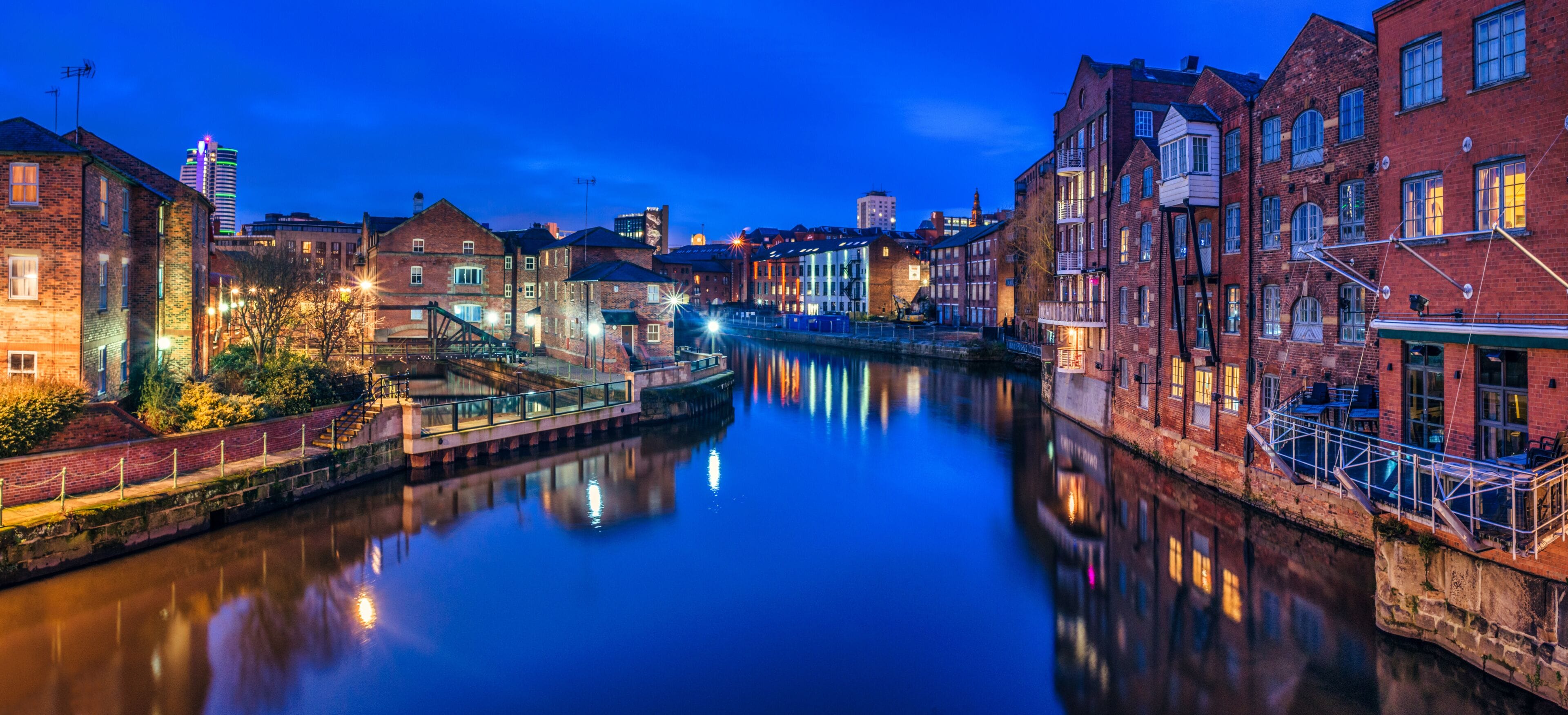 Une rivière paisible où se reflètent des bâtiments, un pont et un gratte-ciel lointain illuminé, au crépuscule.