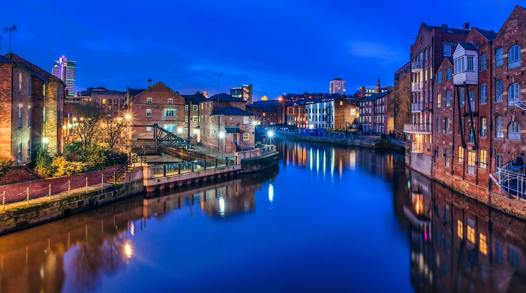 Une rivière paisible où se reflètent des bâtiments, un pont et un gratte-ciel lointain illuminé, au crépuscule.