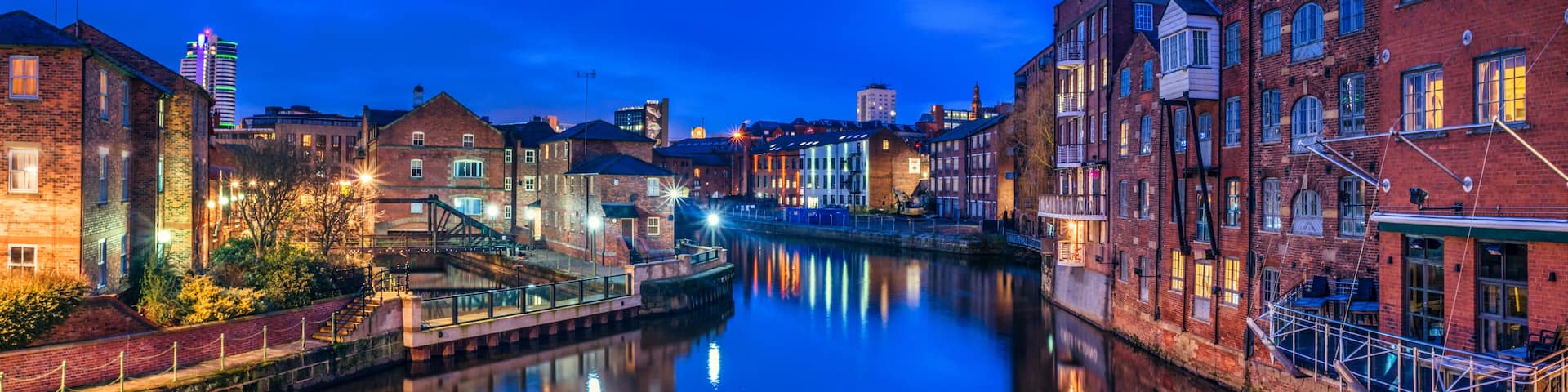 A calm river reflecting buildings, a bridge, and a distant illuminated skyscraper at twilight.