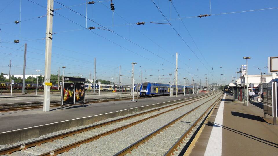 A train station with multiple tracks, overhead electrical lines, and a blue train on the right.