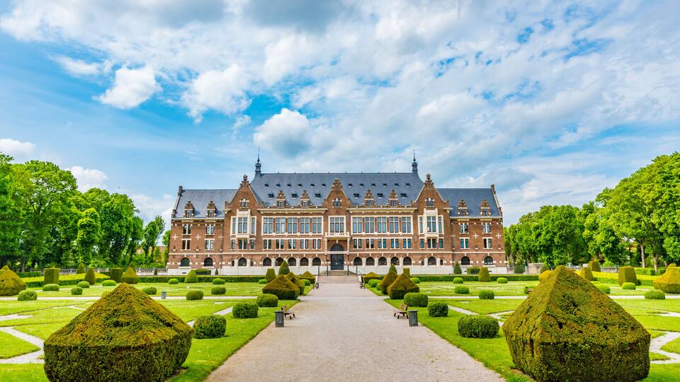 A large, historic building with a symmetrical facade, surrounded by a well-manicured garden with geometrically shaped hedges and a central pathway leading to the entrance.