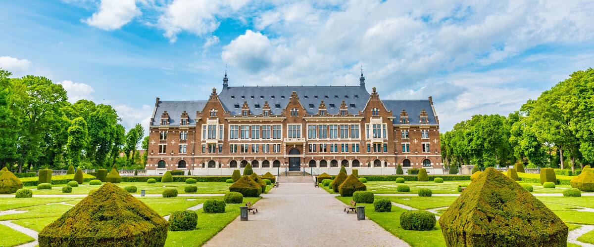A large, historic building with a symmetrical facade, surrounded by a well-manicured garden with geometrically shaped hedges and a central pathway leading to the entrance.