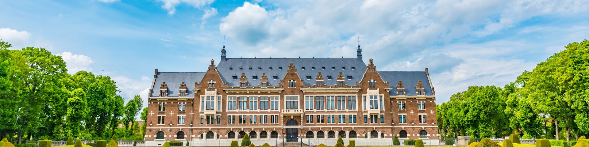 A large, historic building with a symmetrical facade, surrounded by a well-manicured garden with geometrically shaped hedges and a central pathway leading to the entrance.