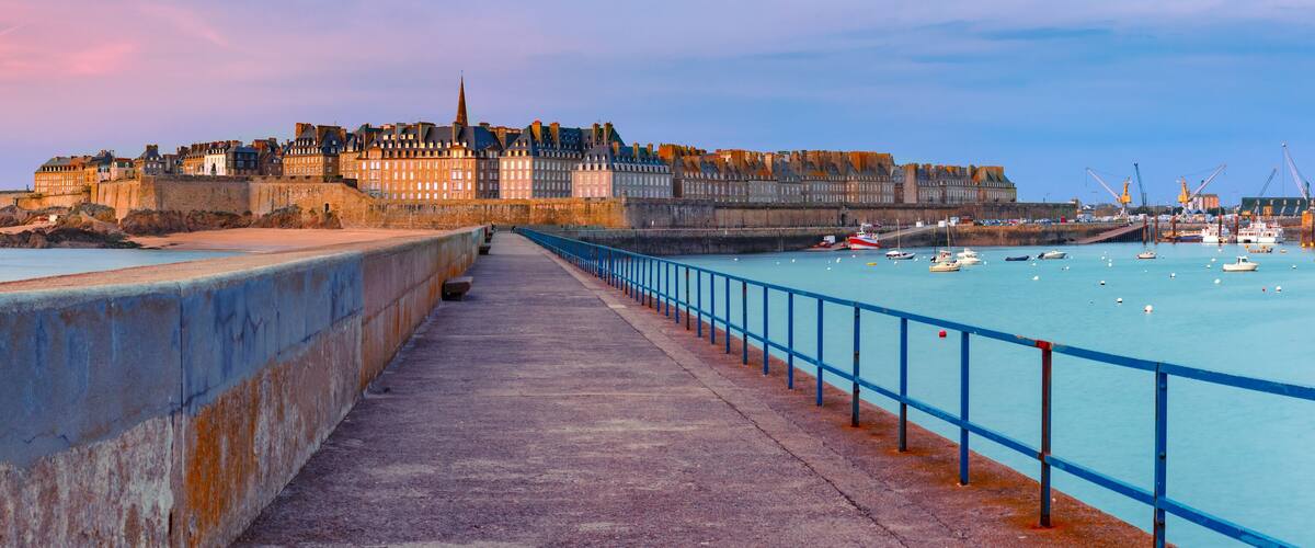 A coastal promenade with a historic castle, a calm sea, and boats.