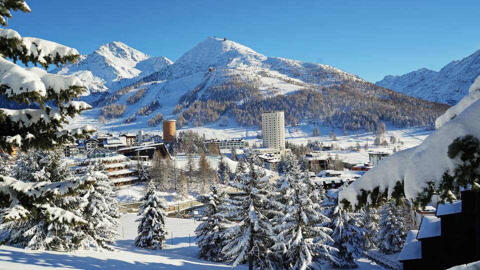 A snowy mountain landscape with a ski resort, buildings, and a clear blue sky.