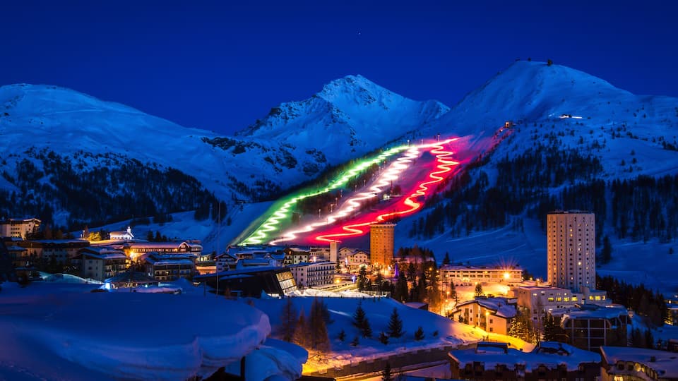 A snow-covered mountain range at dusk with illuminated ski slopes and buildings.