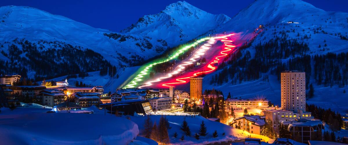 A snow-covered mountain range at dusk with illuminated ski slopes and buildings.