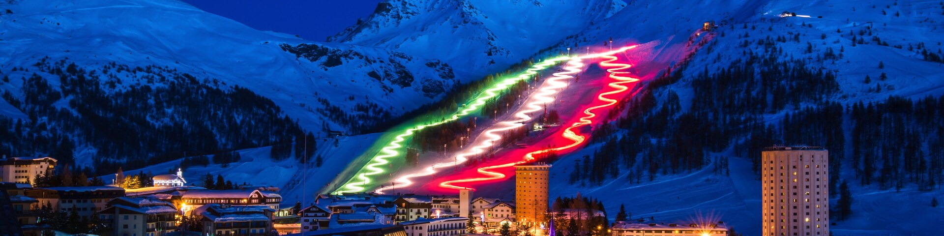 A snow-covered mountain range at dusk with illuminated ski slopes and buildings.