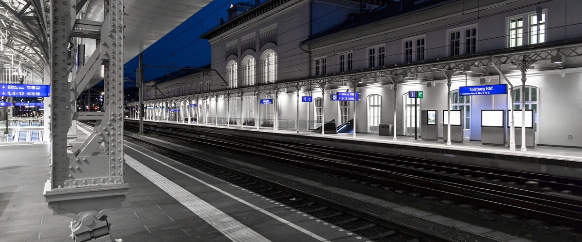Un quai de gare la nuit, avec des voies ferrées, un bâtiment doté de fenêtres en arc de cercle et des panneaux lumineux.