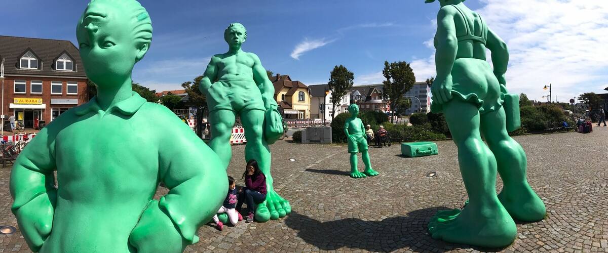 A public square with large green statues, cobblestone pavement, and surrounding buildings.