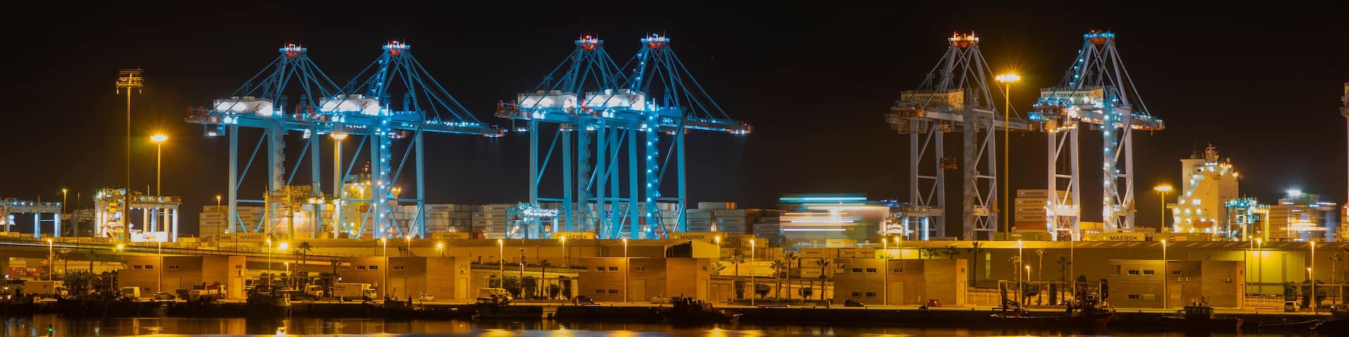 Vista nocturna de un puerto con grúas iluminadas y edificios industriales reflejándose en el agua.