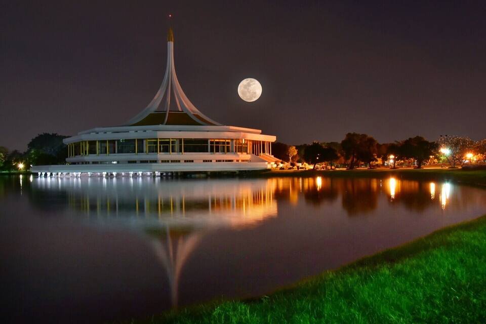 A modern building with a unique spire, reflecting in a calm body of water at night, with a full moon in the sky.