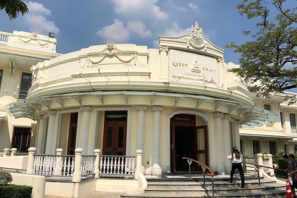A historic building with ornate architecture, including a dome and columns, and a sign reading "Queen Sirikit Museum of Textiles."
