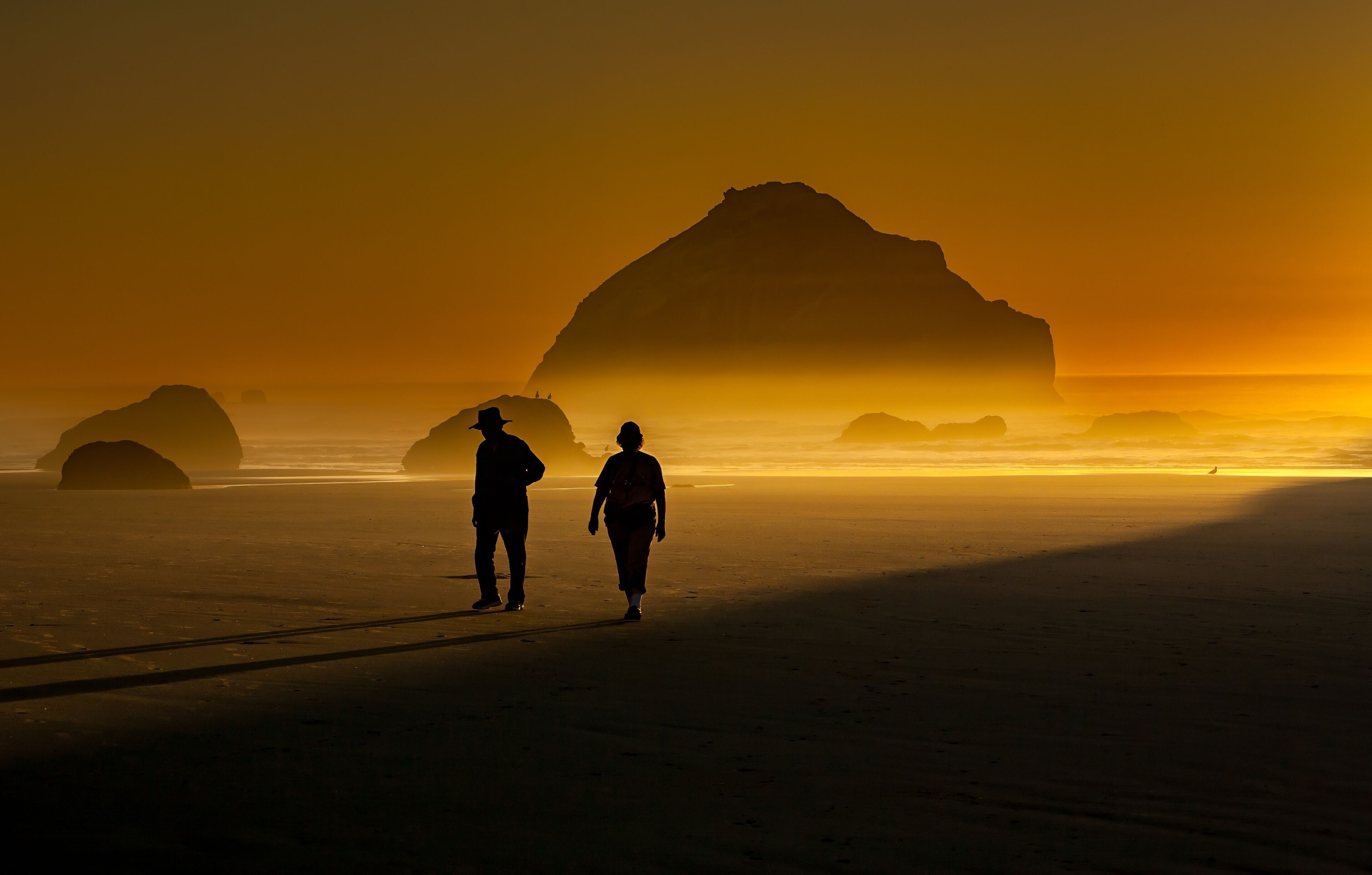 Dos personas caminando por una playa con una gran formación rocosa al fondo durante el atardecer.