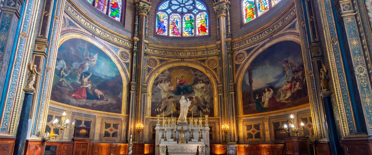 Interior de una iglesia con vidrieras ornamentales, un altar central y frescos detallados en las paredes.