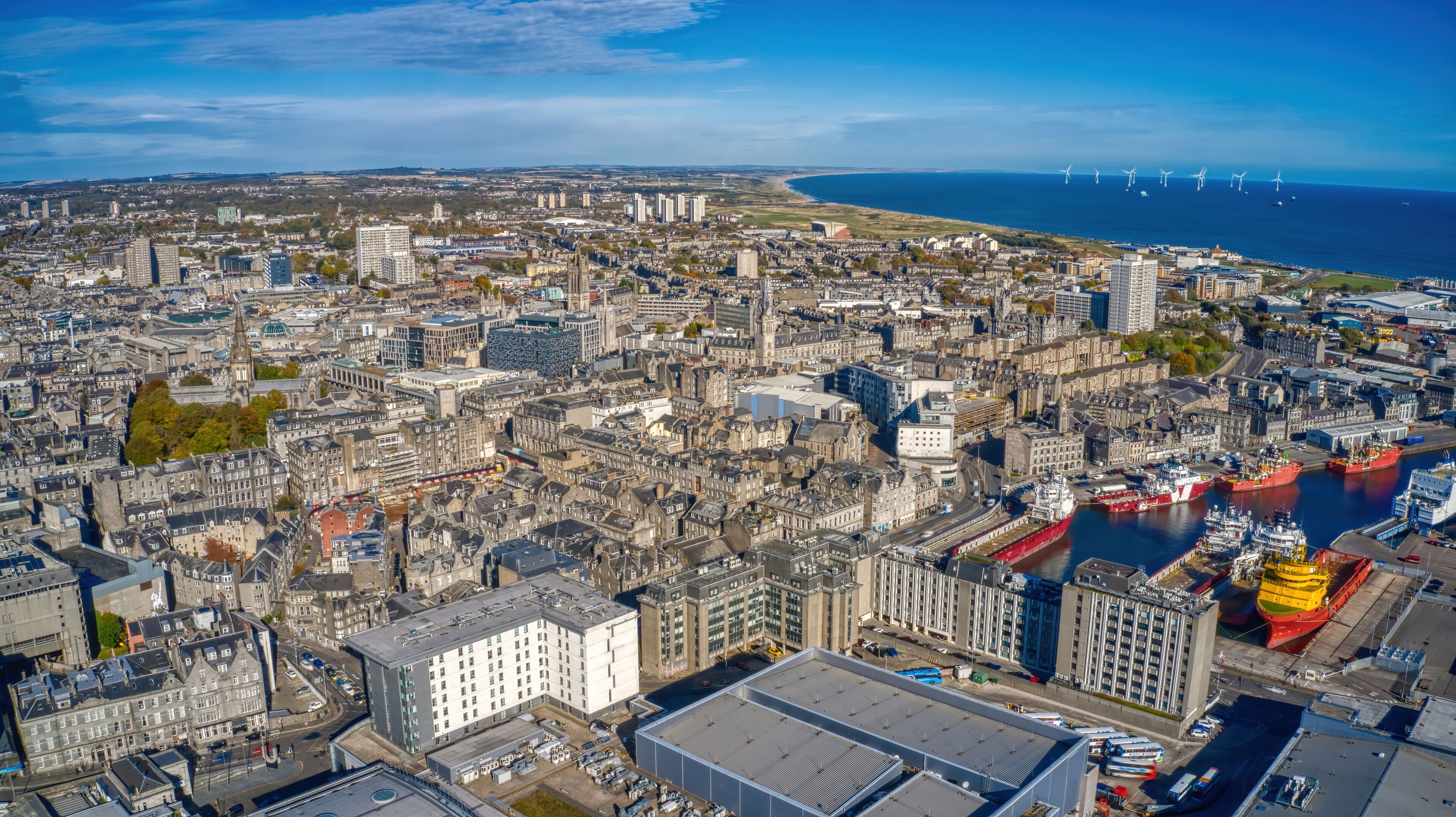 A coastal cityscape with dense urban architecture, a harbor with large ships, and wind turbines in the distance.