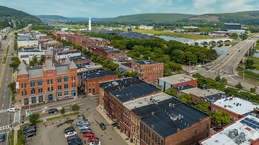 Aerial view of Corning Steuben County, New York downtown, Market Street, glass factory, chemung river, centerway walking bridge, little joe tower, parking lot with cloudy blue sky
