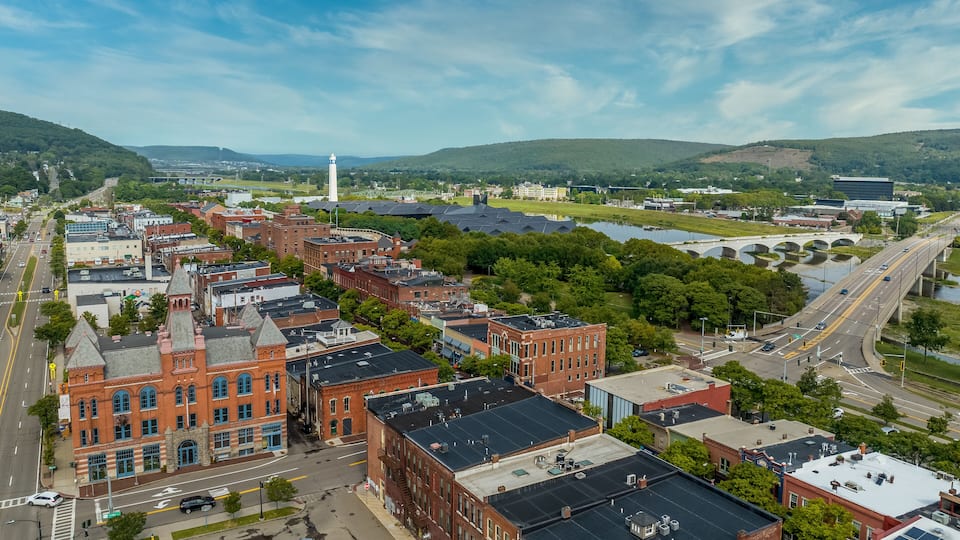 Aerial view of Corning Steuben County, New York downtown, Market Street, glass factory, chemung river, centerway walking bridge, little joe tower, parking lot with cloudy blue sky