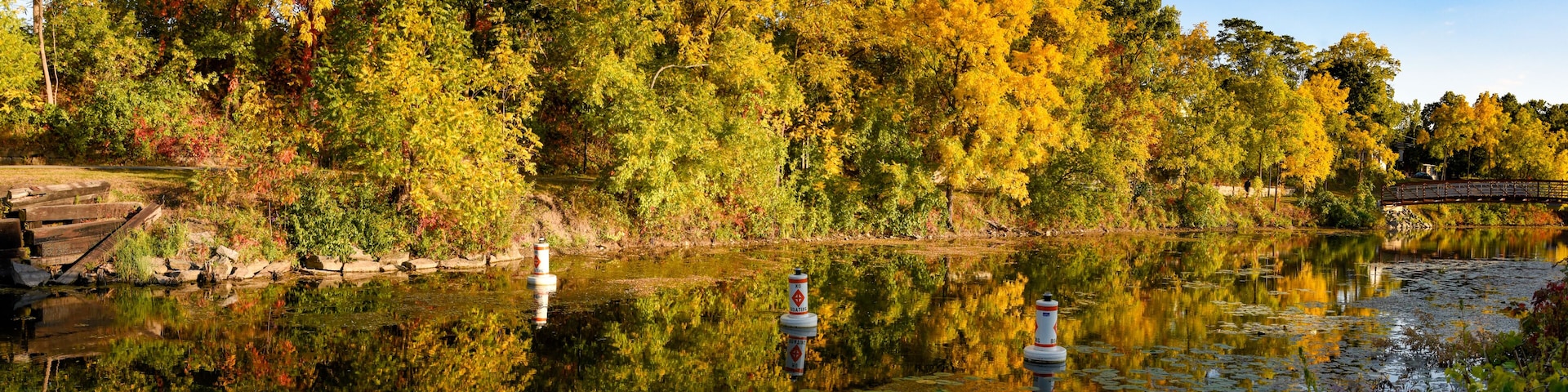 A calm lake with autumn foliage, a wooden dock, and a bridge in the background.