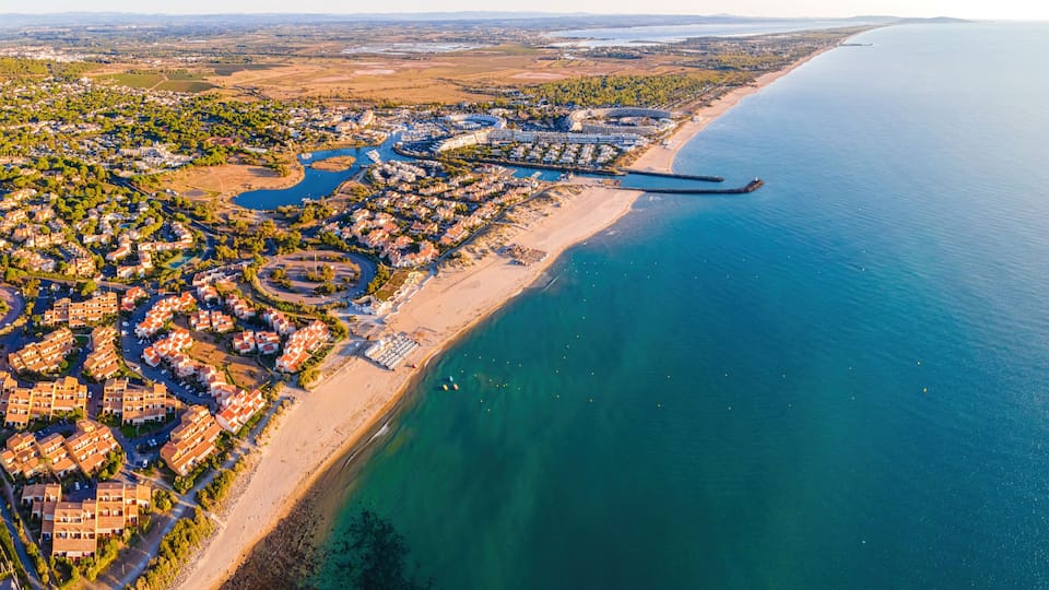 An aerial view of a coastal town with a marina, residential areas, and a long beach.